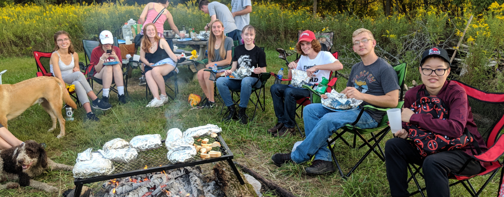  A group of students gathered around a fire cooking and eating food. In the bottom left corner a fire is burning and tin foil-wrapped food is on a grate above the fire.  