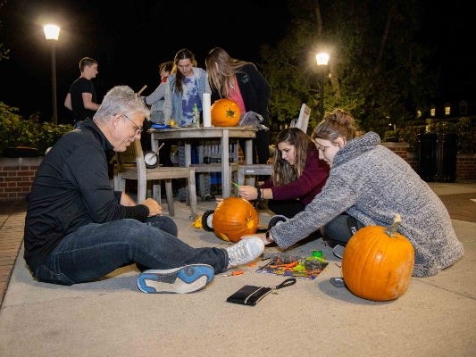  3 people in foreground, sitting on the ground carving pumpkins, 4 in the background standing around a partially completed cart