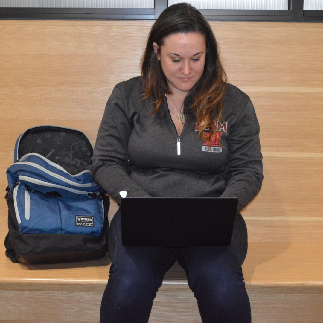 Female student sitting on a bench in the hallway of Shriver near the rinella center, working on a laptop