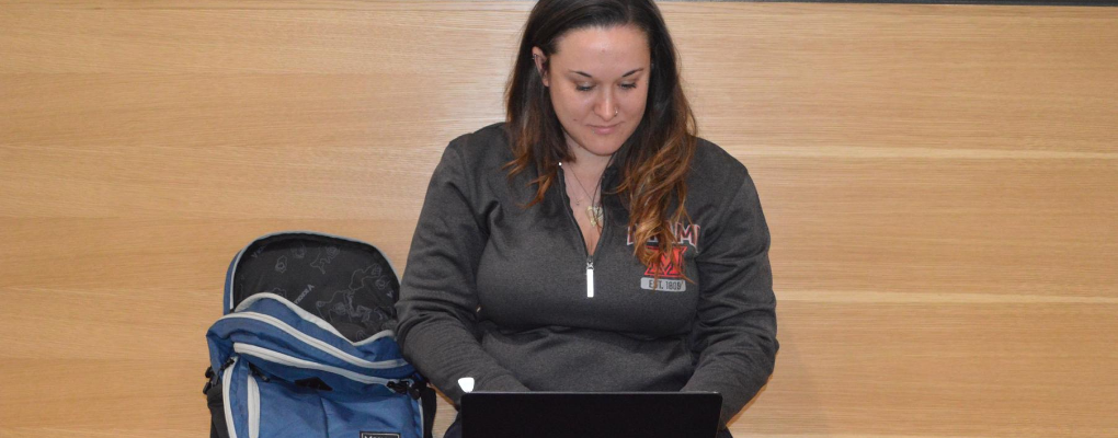 Female student sitting on a bench in the hallway of Shriver near the rinella center, working on a laptop