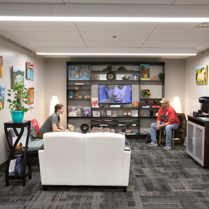 Two students sitting in the Miller Center Lobby, a comfortable and tastefully decorated room with art on the walls, a large bookshelf with knickknacks and art, and a coffee maker and water cooler. 